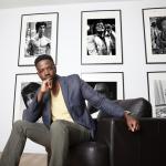 A man in a sportcoat sits in a chair with photos of iconic figures behind him. 