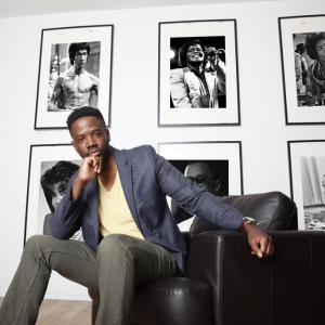 A man in a sportcoat sits in a chair with photos of iconic figures behind him. 