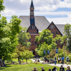 The image shows a vibrant campus scene at Montana State University. Students are walking, sitting, and conversing on a sunny day. In the background, a historic brick building with a tall clock tower and steep gabled roofs is framed by lush green trees. Th