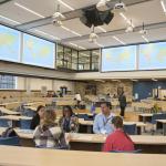 A group of five people sit at a table talking in a large classroom with a world map protected on several screens high in the background