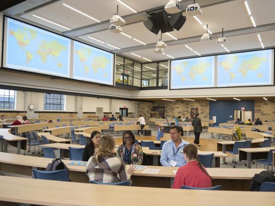 A group of five people sit at a table talking in a large classroom with a world map protected on several screens high in the background | MSU Photo by Colter Peterson