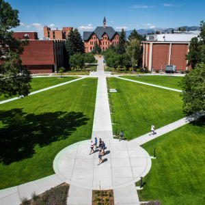 An elevated view of the Montana State University campus shows a group of people walking on a circular concrete pathway that branches out into multiple directions across a large, neatly manicured green lawn. The path leads towards several red-brick buildin