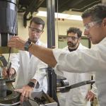 Three men stand around a scientific machine while two of them reach in to adjust something on the machine