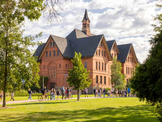 The image shows a large group of students walking and biking across the green, tree-lined lawn of Montana State University. In the background is the iconic brick structure of Montana Hall, with its steep gabled roofs and central cupola. The sky is partly  | Marcus "Doc" Cravens/MSU Photo