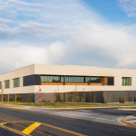 A modern two-story commercial building with a white and gray exterior sits at the corner of an urban street under a blue, partly cloudy sky. Large windows line the upper and lower levels, and the building features clean architectural lines with landscaped