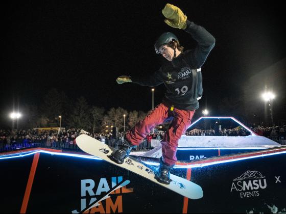 A person on a snowboard slides on a metal rail with a large crowd of spectators in the background | MSU Photo by Colter Peterson