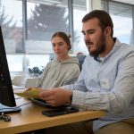  male student wearing a light blue button-up shirt and a name tag reading "Josh" sits at a computer station in a classroom, holding a yellow sheet of paper. A female student in a gray hoodie sits beside him, attentively looking at the paper. Other