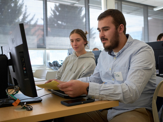  male student wearing a light blue button-up shirt and a name tag reading "Josh" sits at a computer station in a classroom, holding a yellow sheet of paper. A female student in a gray hoodie sits beside him, attentively looking at the paper. Other | Kelly Gorham/MSU