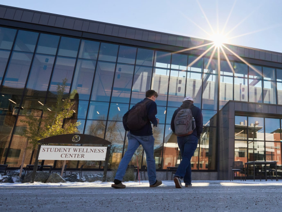 Two students walk toward the Student Wellness Center at Montana State University, a long building with many large windows. The sun peaks over the top of the building. Light snow dusts the ground. | Colter Peterson/MSU