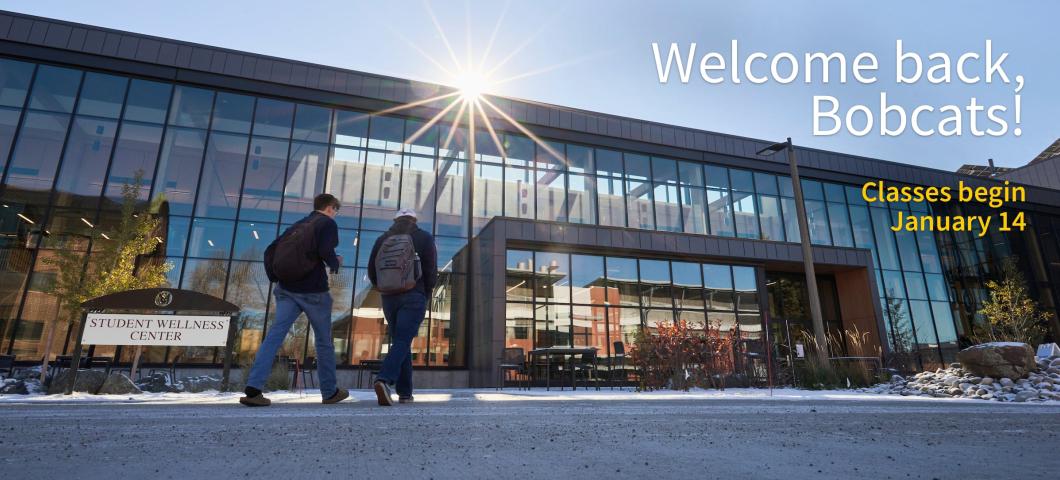 Two students walk toward the Student Wellness Center at Montana State University, a long building with many large windows. The sun peaks over the top of the building. Light snow dusts the ground. | Colter Peterson/MSU