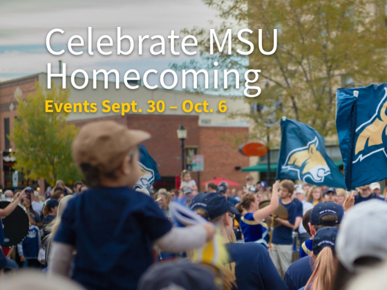 The image shows a vibrant scene from an outdoor event celebrating Montana State University's Homecoming. A large crowd of people is gathered in a downtown area, some holding blue flags featuring the MSU Bobcat logo. In the foreground, a small child is | MSU Image