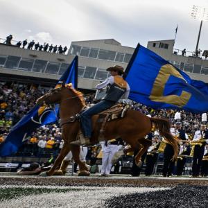 Riders on horseback carry large blue flags with gold designs across a football field, leading a marching band dressed in gold and white uniforms. In the background, a packed stadium with fans and a prominent university building is visible under a partly c