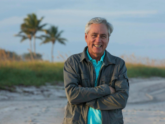 A smiling man with short gray hair stands on a sandy beach with his arms crossed. He is wearing a teal polo shirt under a light gray jacket. In the background, palm trees and greenery are visible under a clear sky. | 