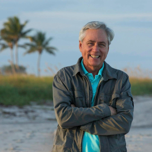 A smiling man with short gray hair stands on a sandy beach with his arms crossed. He is wearing a teal polo shirt under a light gray jacket. In the background, palm trees and greenery are visible under a clear sky.