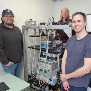 Three men stand around a large technology array while looking at the camera