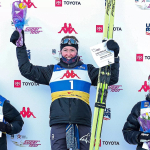 Three female cross-country skiers stand on a podium holding certificates, flowers, and skis in front of a backdrop with sponsor logos including Toyota, US Ski Team, and Super Tour. The central skier, wearing bib number 1, raises her arms in celebration, h