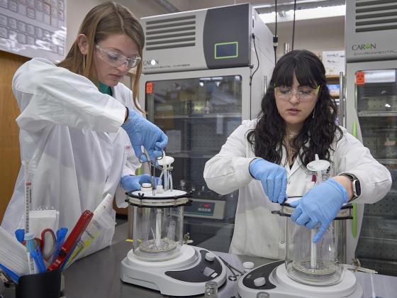 Kylie Bodie and Ghazal Vahid wearing lab jackets and safety equipment working with devices in lab. | MSU photo by Kelly Gorham