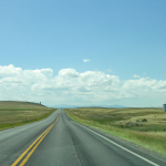 A country road with grasslands on either side and three silos off in the distance. 