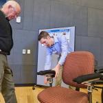 A man watches on the left side of the frame as another man bends over an office chair adjusting mechanical ergonomic arm-rests attached to the chair