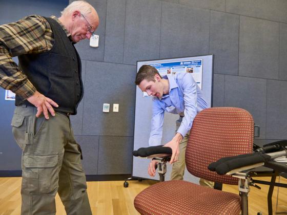 A man watches on the left side of the frame as another man bends over an office chair adjusting mechanical ergonomic arm-rests attached to the chair | MSU photo by Colter Peterson