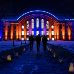 The image shows Romney Hall at Montana State University illuminated at night with blue, purple, and orange lights. The pathway leading to the building is lined with glowing paper lanterns, creating a warm, inviting atmosphere against the snow-covered grou