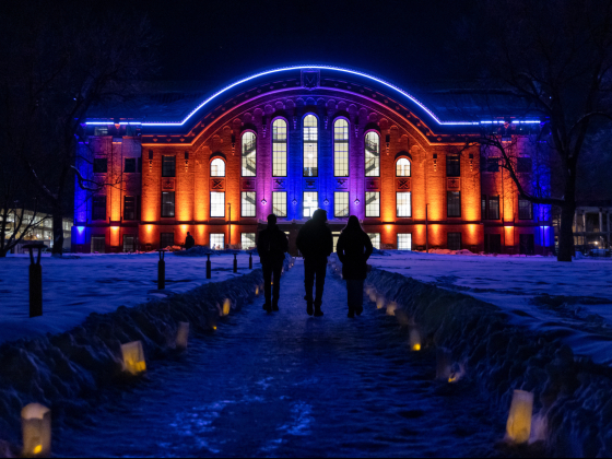 The image shows Romney Hall at Montana State University illuminated at night with blue, purple, and orange lights. The pathway leading to the building is lined with glowing paper lanterns, creating a warm, inviting atmosphere against the snow-covered grou | Marcus "Doc" Cravens/MSU