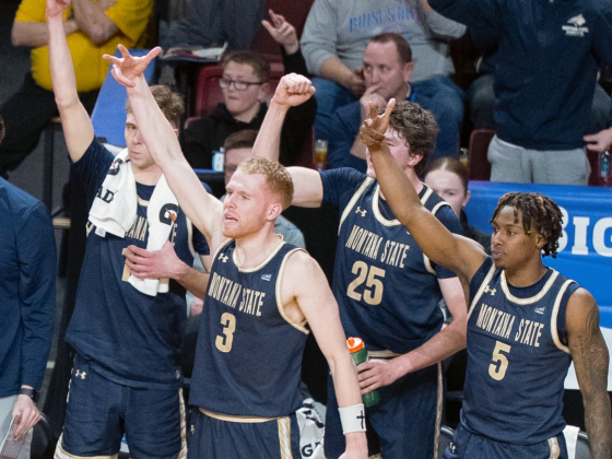 A promotional graphic featuring Montana State University men’s basketball players celebrating on the sidelines, wearing navy blue uniforms with gold lettering. Players are raising their arms and cheering, with some standing and others seated. A referee in | Image from Bobcat Athletics 