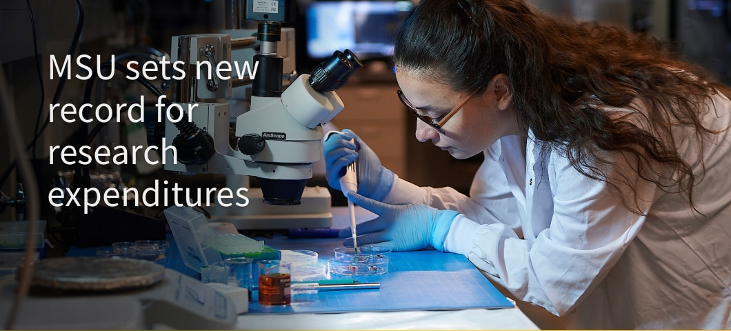 The image shows a researcher wearing a white lab coat and blue gloves working in a laboratory. She is using a pipette to place a sample into a petri dish while looking through a microscope. The laboratory setting is equipped with advanced scientific tools | MSU Image