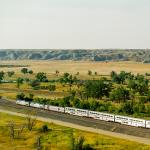 The image shows a scenic landscape in northern Montana with a long passenger train traveling through a grassy plain. The foreground is dotted with green trees and fields, while the background features rugged, rocky hills under a hazy sky.