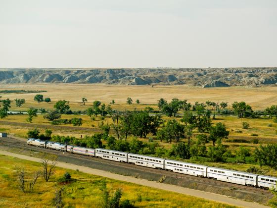 The image shows a scenic landscape in northern Montana with a long passenger train traveling through a grassy plain. The foreground is dotted with green trees and fields, while the background features rugged, rocky hills under a hazy sky. | Kelly Gorham/Montana State Universityi