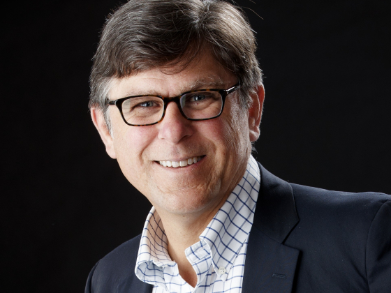 The image shows a man with short, dark hair and glasses, smiling while looking at the camera. He is wearing a dark blazer over a white shirt with a blue checkered pattern. The background is black, which contrasts with his light-colored clothing and makes  | 