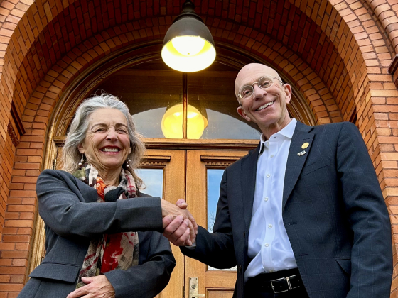 A woman and a man stand on the steps of a historic red-brick building, shaking hands and smiling at the camer. The woman wears a dark gray suit with a floral scarf, while the man wears a dark blazer, white shirt, and a lapel pin. The arched brick entrance | Photo by Todd Wilkinson