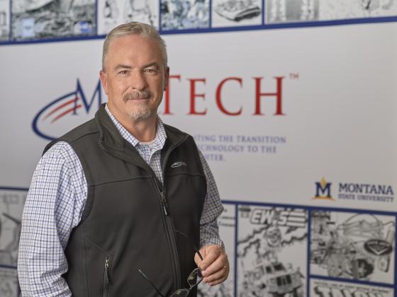 a man in a collard shirt and vest stands, looking at the camera, in front of a large banner reading MilTech | MSU photo by Colter Peterson