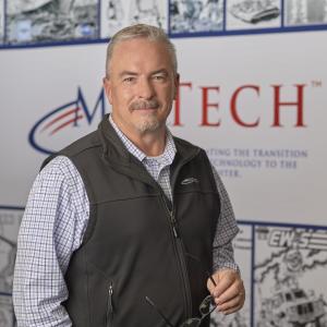 a man in a collard shirt and vest stands, looking at the camera, in front of a large banner reading MilTech