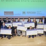 A group of people is standing on a stage at Montana State University, posing for a photo while holding large ceremonial checks. The backdrop features logos for "Blackstone LaunchPad," "Montana State University," and the "Jake Jabs Coll