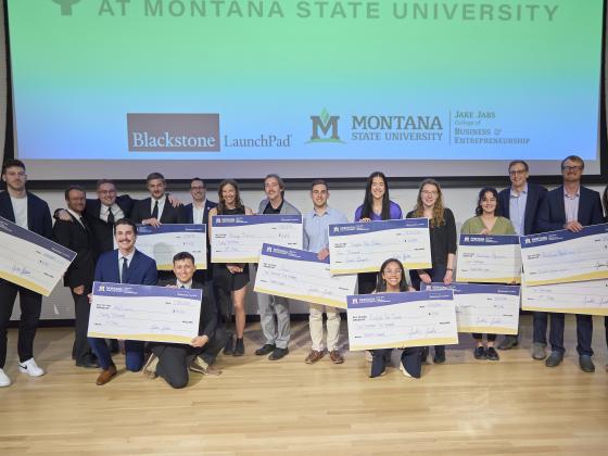 A group of people is standing on a stage at Montana State University, posing for a photo while holding large ceremonial checks. The backdrop features logos for "Blackstone LaunchPad," "Montana State University," and the "Jake Jabs Coll | Colter Peterson