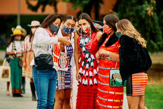 American Indian students wearing fashion dresses and fashion masks huddle as they look at photos on one of the students’ phones.