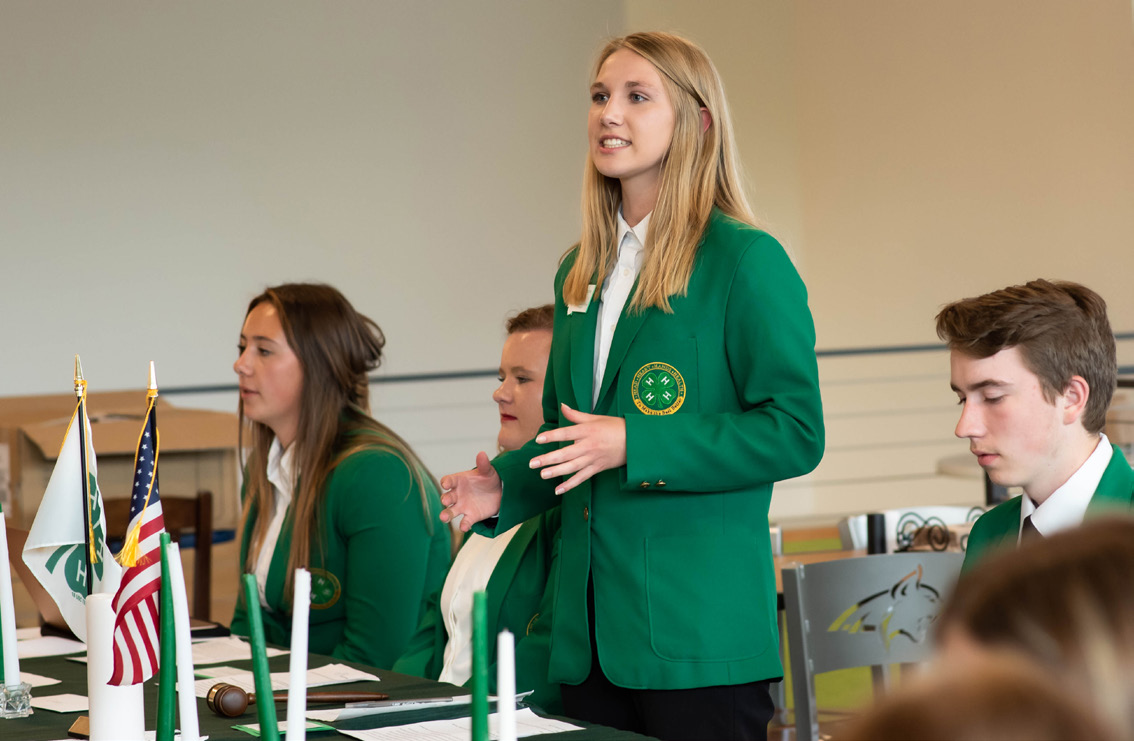 A girl is speaking amongst a group of 4-H leaders. They all have on green 4-H blazers.