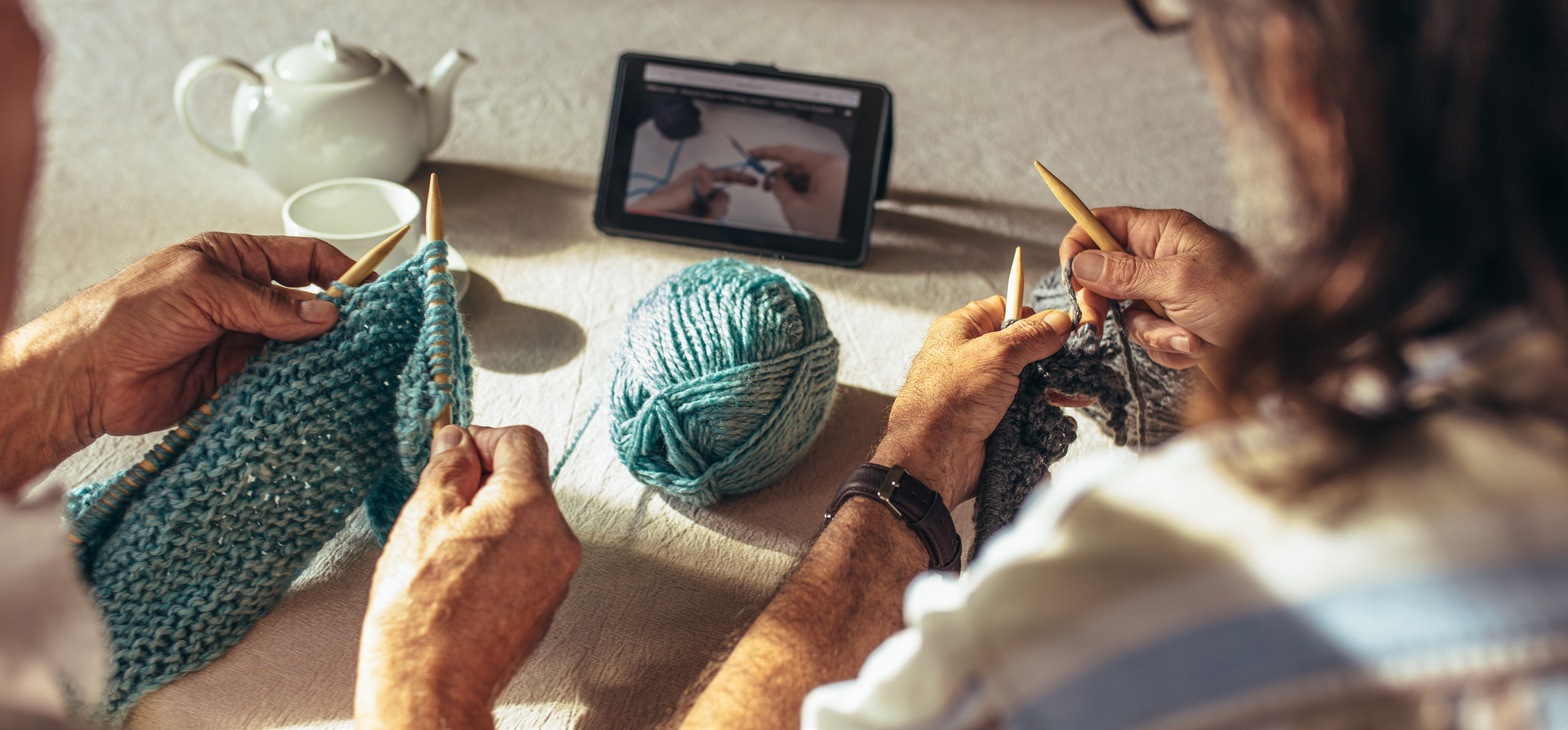 Two people watch a tutorial video on how to knit as they hold knitting needles and blue yarn in their hands.