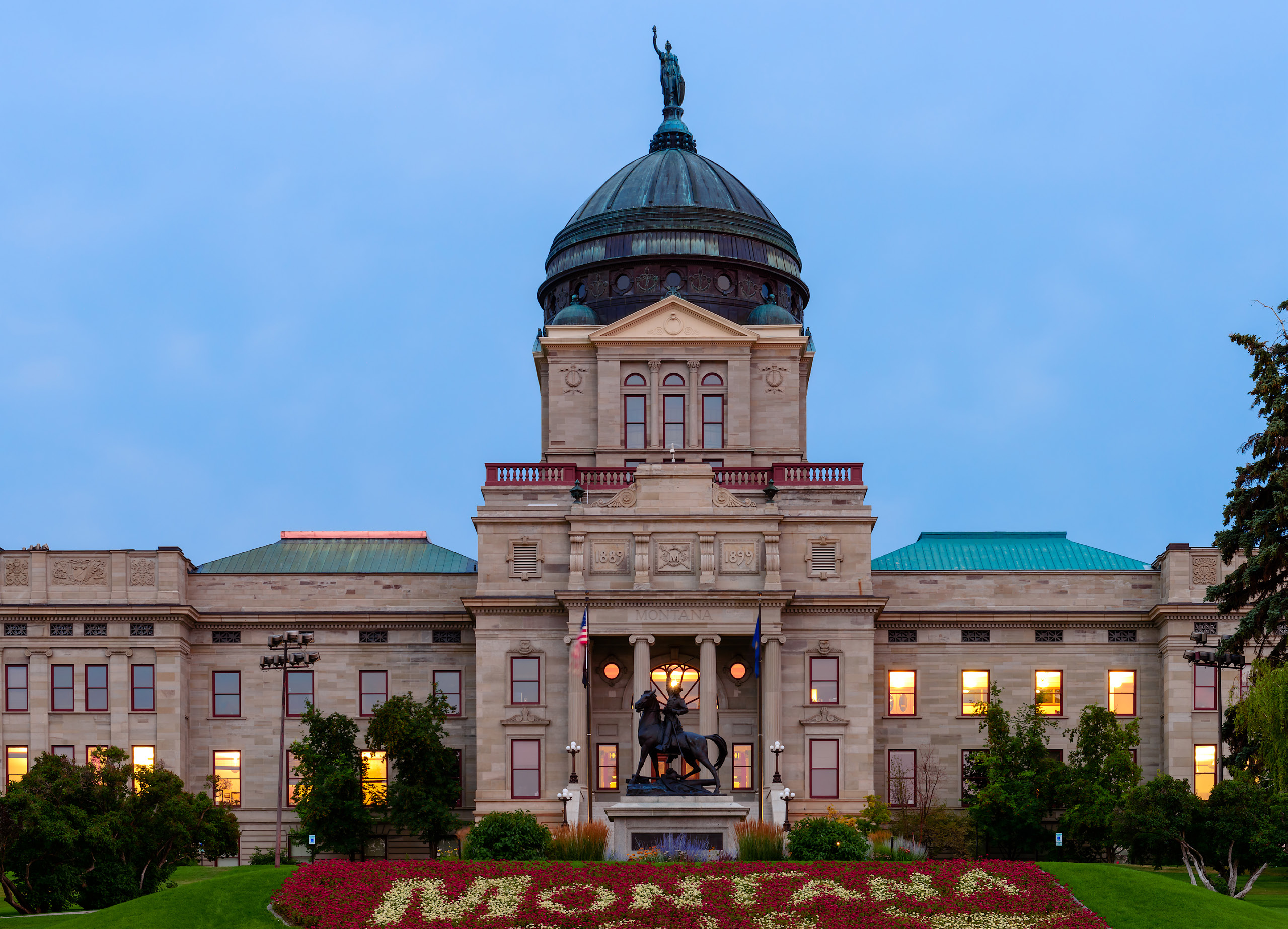 Image of Montana's capitol building at dusk with a light blue sky in the background and windows lit. There is a horse statute in front with a well tended lawn. The building rises tall with a small statue of a human at the very top.