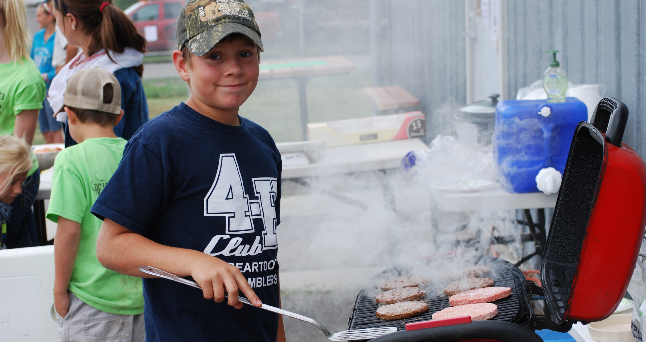 Young boy grilling hamburger patties on a red grill with a silver spatula in hand.