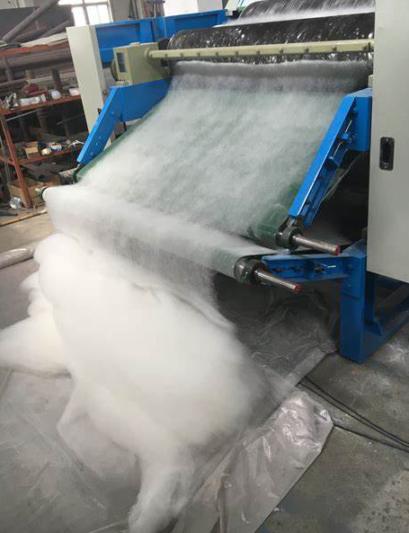 A blue carding machine aligns white wool fibers through metal rollers.