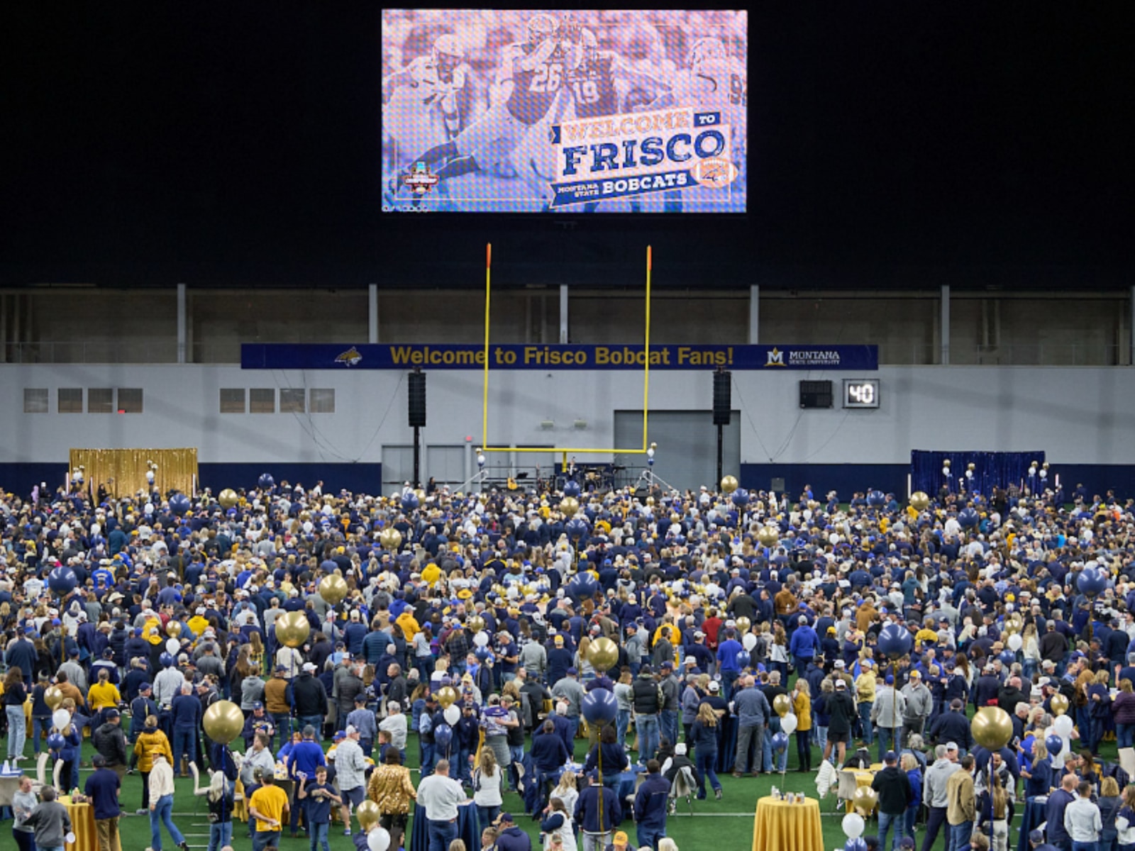 Image of a large crowd of Montana State University supports on the field at the Dallas Cowboys' practice facility in Frisco, Texas, for an event ahead of the national championship game. A large video board shows a still from a football game, and the message "Welcome to Brisco Bobcat Fans" appears on banners.