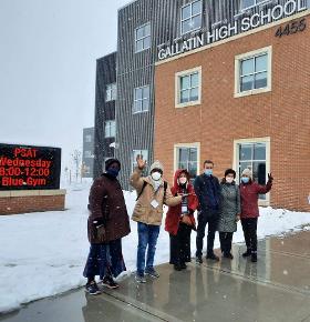 6 people look incredibly cold as they pose with excitement in the snow, in front of a large building.