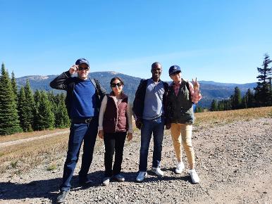 4 people pose at the top of a hike, pine trees in the background. It looks sunny and the sky is blue. 