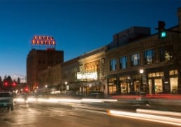 A long exposure shot at night shows streaked headlights of cars moving down a city street. Colorfully lit buildings are in the background.