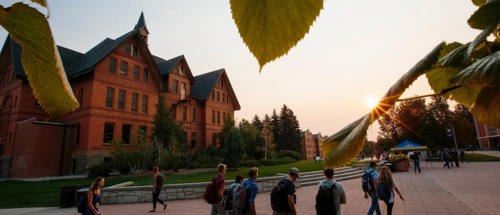 Students walk along a mall at the Montana State University campus as the sun rises over academic buildings in the background.