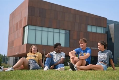 Four students sit on the grass at a college campus with an angular, modern building in the background on a sunny day.