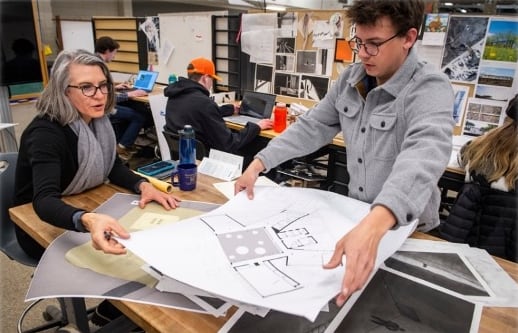 A professor works with a student over a table full of papers and diagrams in a studio environment. 