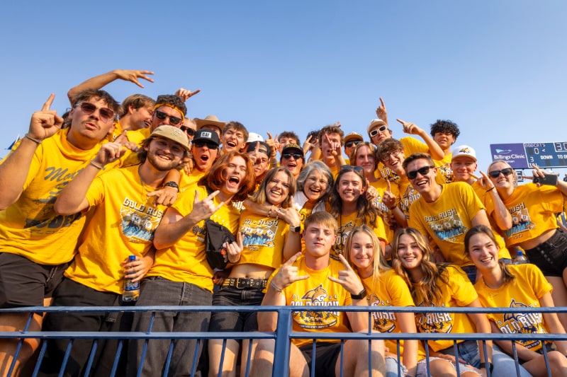 The image shows a large group of enthusiastic Montana State University students and fans wearing matching gold shirts that say "Gold Rush." They are packed closely together in the stands of a stadium, smiling, cheering, and making celebratory gestures like holding up their fingers to signify "number one."